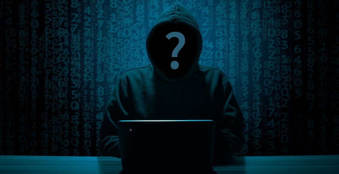 Zakapturzona postać z pytajnikiem zamiast twarzy siedzi przed laptopem w ciemnoniebieskim pokoju.