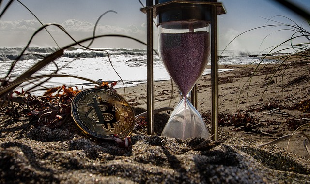 Bitcoin leżący na plaży, obok klepsydra