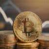 Bitcoin leżący na stosie monet, w tle wykres giełdowy.