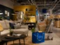 Wnętrze nowoczesnego sklepu meblowego IKEA z urządzonym salonem, sofą, stolikami i wózkiem wypełnionym paczkami oraz zwiniętymi dywanami.