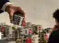 Makieta osiedla mieszkaniowego z czerwonymi metkami symbolizującymi rabaty. Dłoń dewelopera trzyma miniaturowy budynek, a potencjalny kupujący przygląda się dostępnym nieruchomościom.