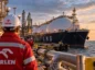 Pracownicy Orlenu w czerwonych kombinezonach obserwują ogromny gazowiec LNG zacumowany przy terminalu przeładunkowym.