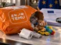 Pomarańczowa przesyłka Temu z zabawkami dziecięcymi i ładowarką USB na marmurowym blacie. W tle flaga Unii Europejskiej i dokumenty dotyczące kontroli bezpieczeństwa produktów.