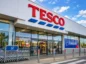 Wejście do sklepu Tesco z charakterystycznym czerwonym logo i rzędami wózków zakupowych przed budynkiem. Grafika ilustruje sprzedaż działalności Tesco w Czechach i na Słowacji holenderskiej grupie Ahold Delhaize.